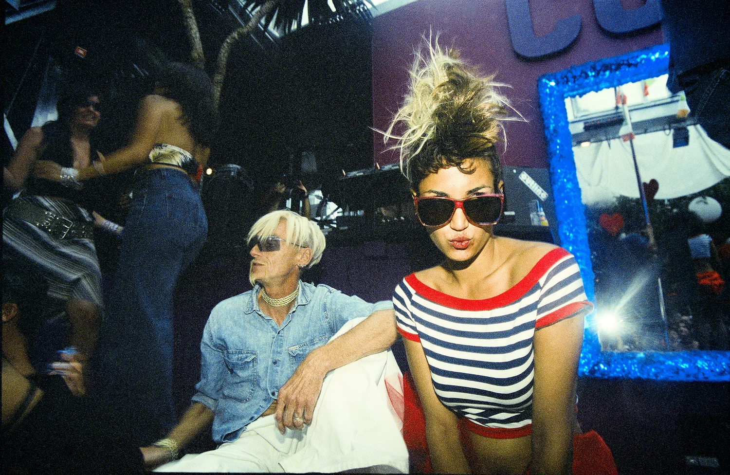 Manumission, Privilege, Ibiza 2001. A clubber in red sunglasses and a striped top beside a glittering blue mirror, a platinum haired clubber in denim behind. 35mm photograph by Brendan Duffy.