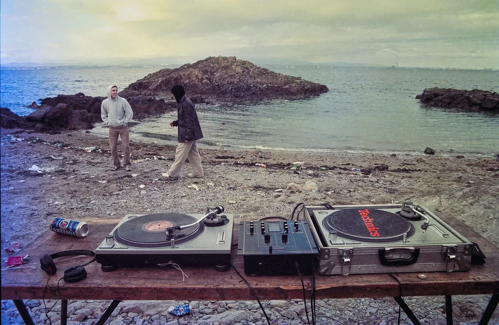 Beach Party, Sutton, Co. Dublin 1999. Two turntables and a mixer on a trestle table on the shore, the sea and rocks behind. 35mm photograph by Brendan Duffy.