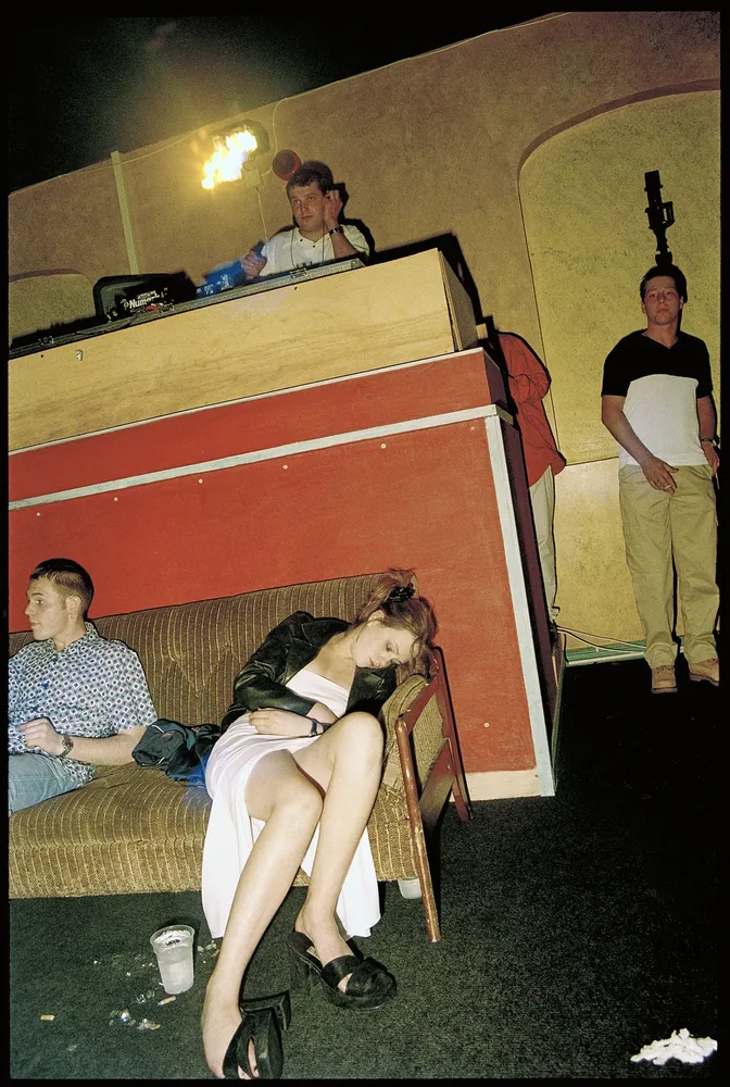 Club Oz, Dungannon, Co. Tyrone 1998. A clubber asleep on a sofa below the DJ booth while the night goes on around them. 35mm photograph by Brendan Duffy.